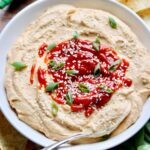 Close up side view of a bowl of hummus topped with bright red sriracha, sesame seeds, and sliced green onions.