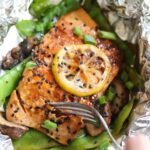 A foil packet of salmon topped with black sesame seeds and a lemon slice, open with a fork taking a bite.