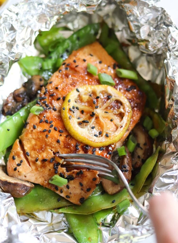 Close up of a foil packet of salmon topped with black sesame seeds and a lemon slice, open with a fork taking a bite.
