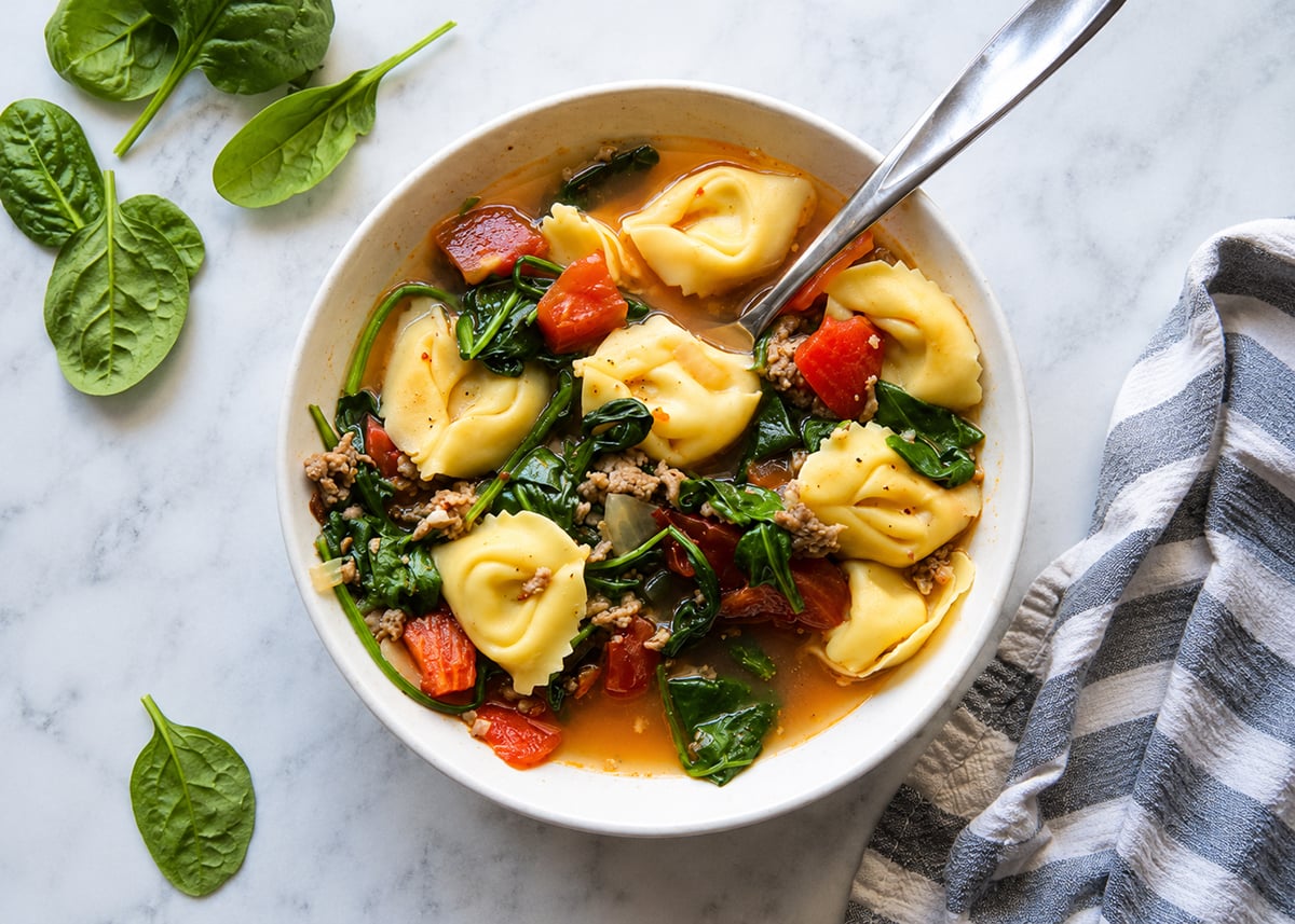 Overhead view of sausage tortellini soup and a spoon in a white soup bowl.