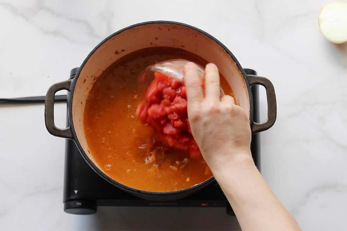 Woman's hand holding a small bowl of diced tomatoes, pouring them into a pot with broth.