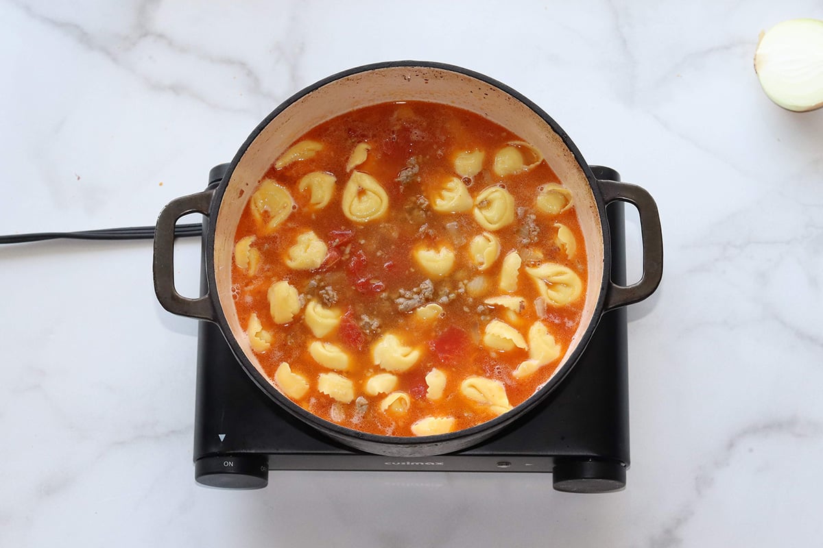 A pot of tortellini soup with tomato broth and ground sausage simmers in a Dutch oven sitting on a black electric hot plate.