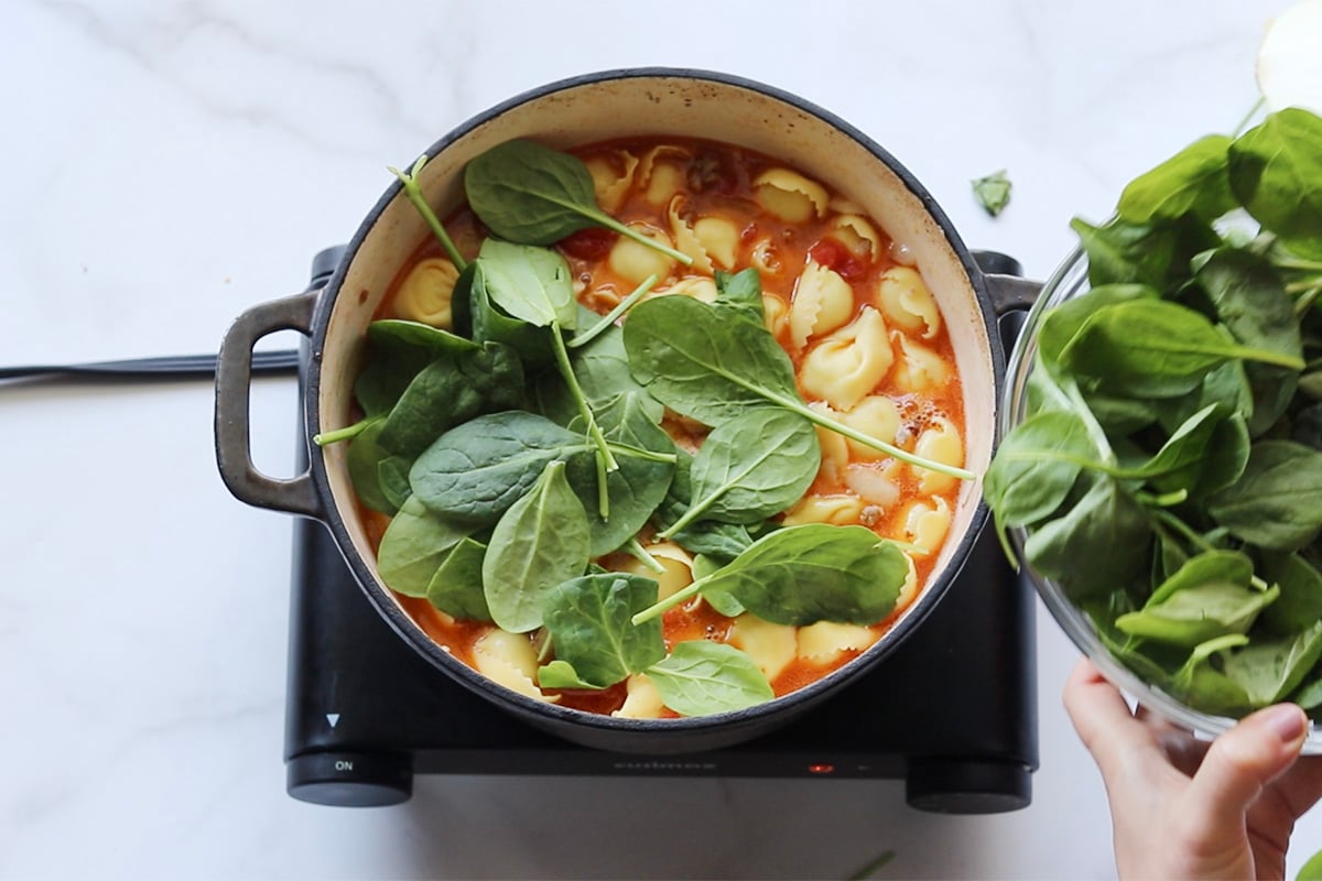 Adding fresh baby spinach to a pot of sausage and tortellini soup, viewed from overhead.