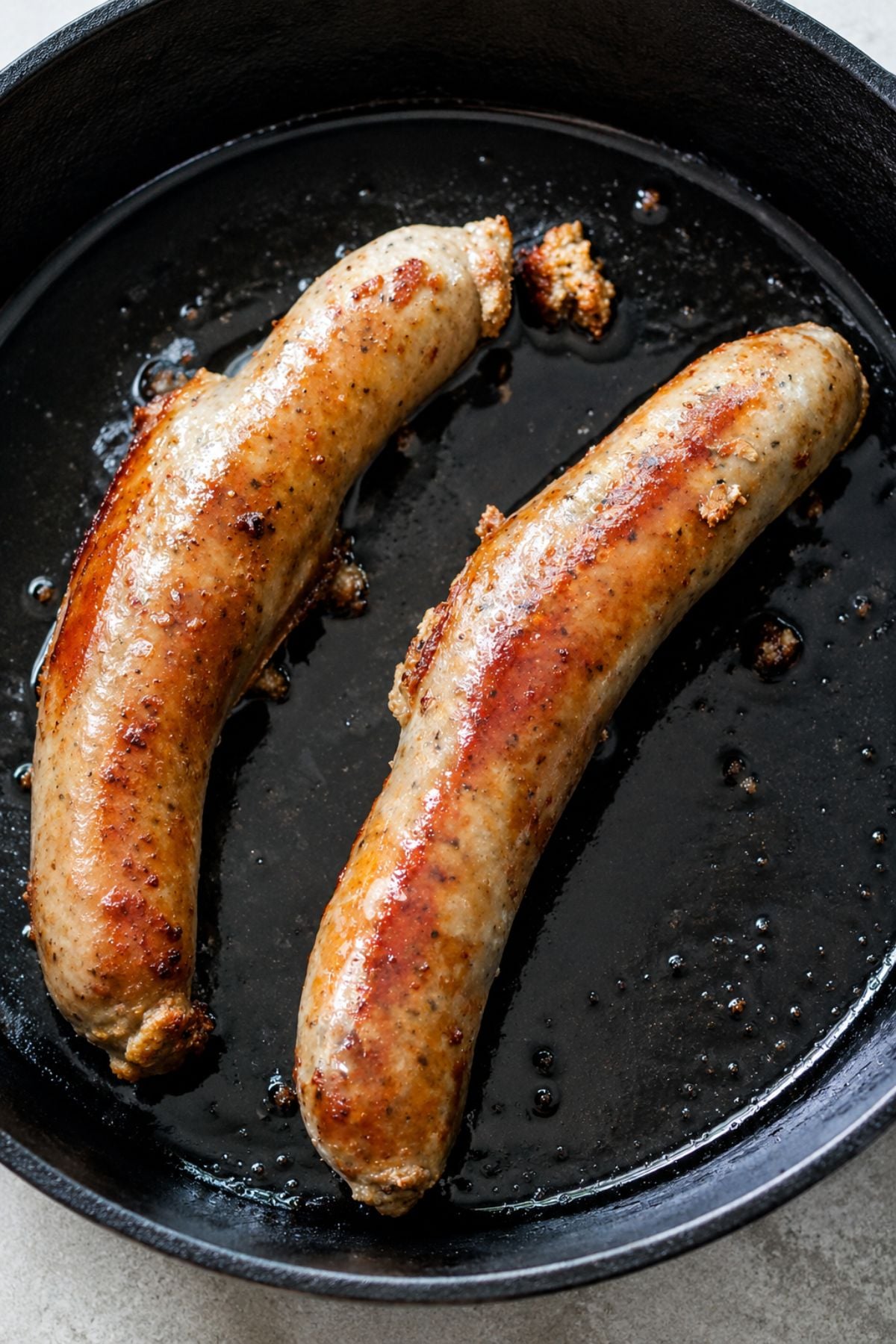 Two browned sausages sizzle in a black cast iron skillet, surrounded by glistening oil and savory browned bits.