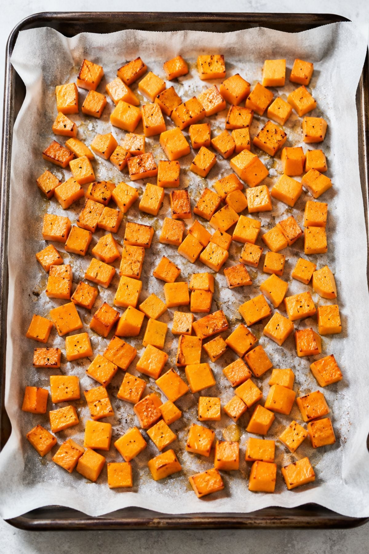Cubed butternut squash, seasoned and roasted, spread evenly on a parchment-lined baking sheet.