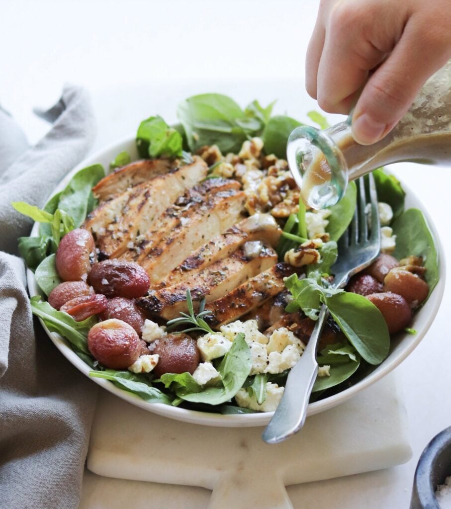 Finished Dish. A hand pours salad dressing from a glass bottle onto a spinach salad in a bowl. The salad is topped with sliced grilled chicken, roasted grapes, crumbled goat cheese, and walnuts.