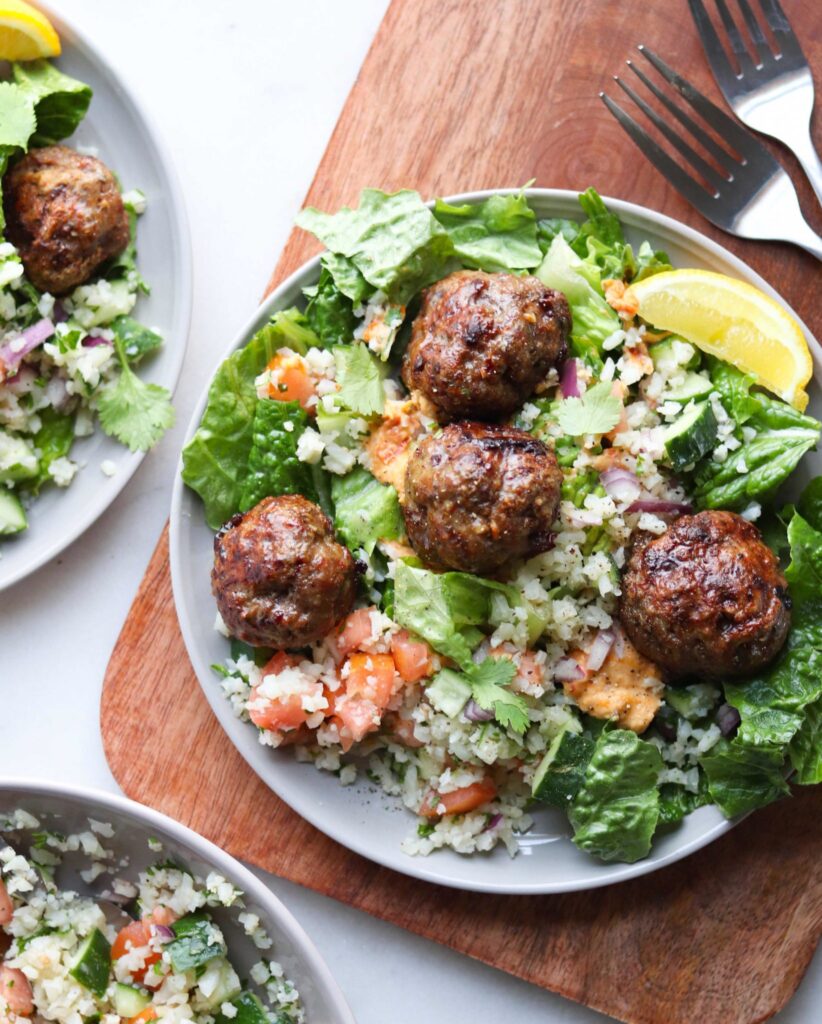Whole30 Mediterranean Lamb Meatballs are served on a gray plate with torn romaine lettuce, cauliflower tabbouleh, hummus and slices of lemon.