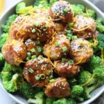 Paleo Orange Chicken Meatballs served over steamed broccoli in a large gray bowl on a white marble board. The meatballs are sprinkled with sliced green onion, orange zest, and sesame seeds.