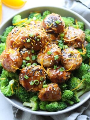 Paleo Orange Chicken Meatballs served over steamed broccoli in a large gray bowl on a white marble board. The meatballs are sprinkled with sliced green onion, orange zest, and sesame seeds.