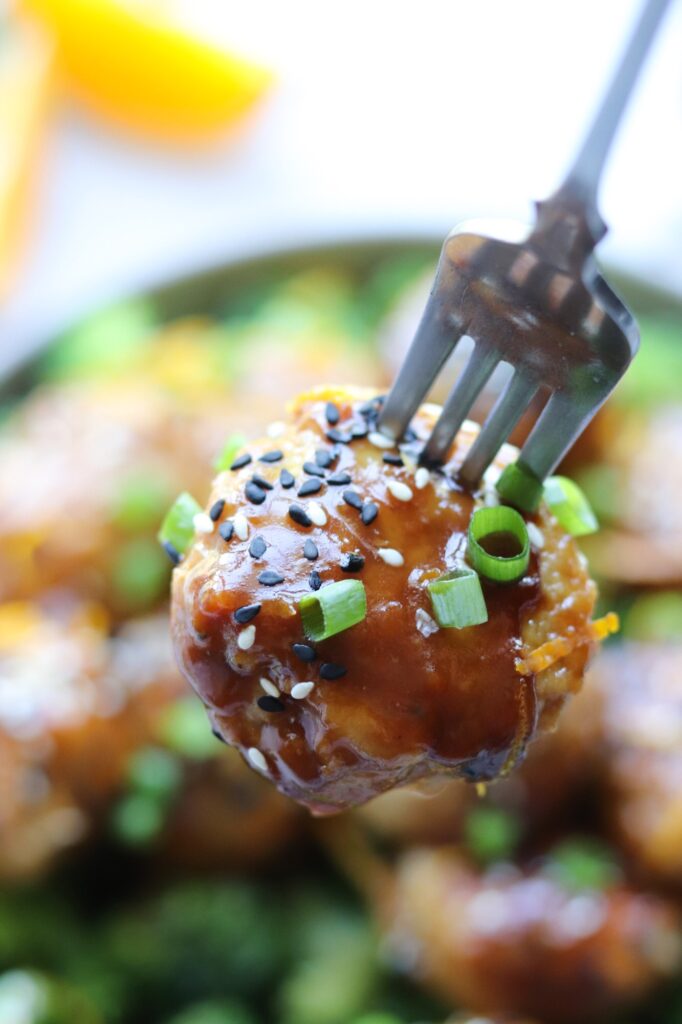 Close up of one glazed meatball on a fork, garnished with sliced green onion, sesame seeds, or orange zest.