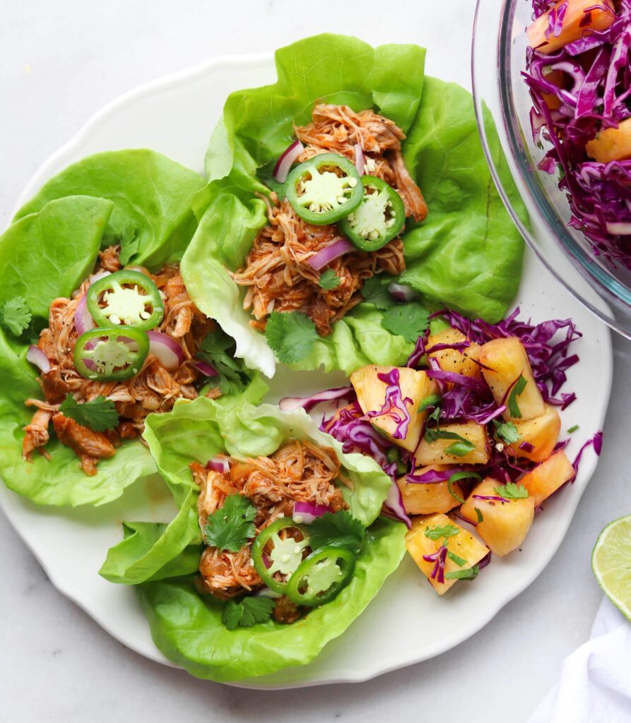 A white plate with three lettuce cups filled with bbq chicken and topped with sliced jalapeno. A hand squeezes a lime on top.