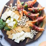 A wooden cutting board lays on a white marble board topped with prosciutto wrapped melon, sliced cheddar cheese, pistachios, and gluten free crackers.