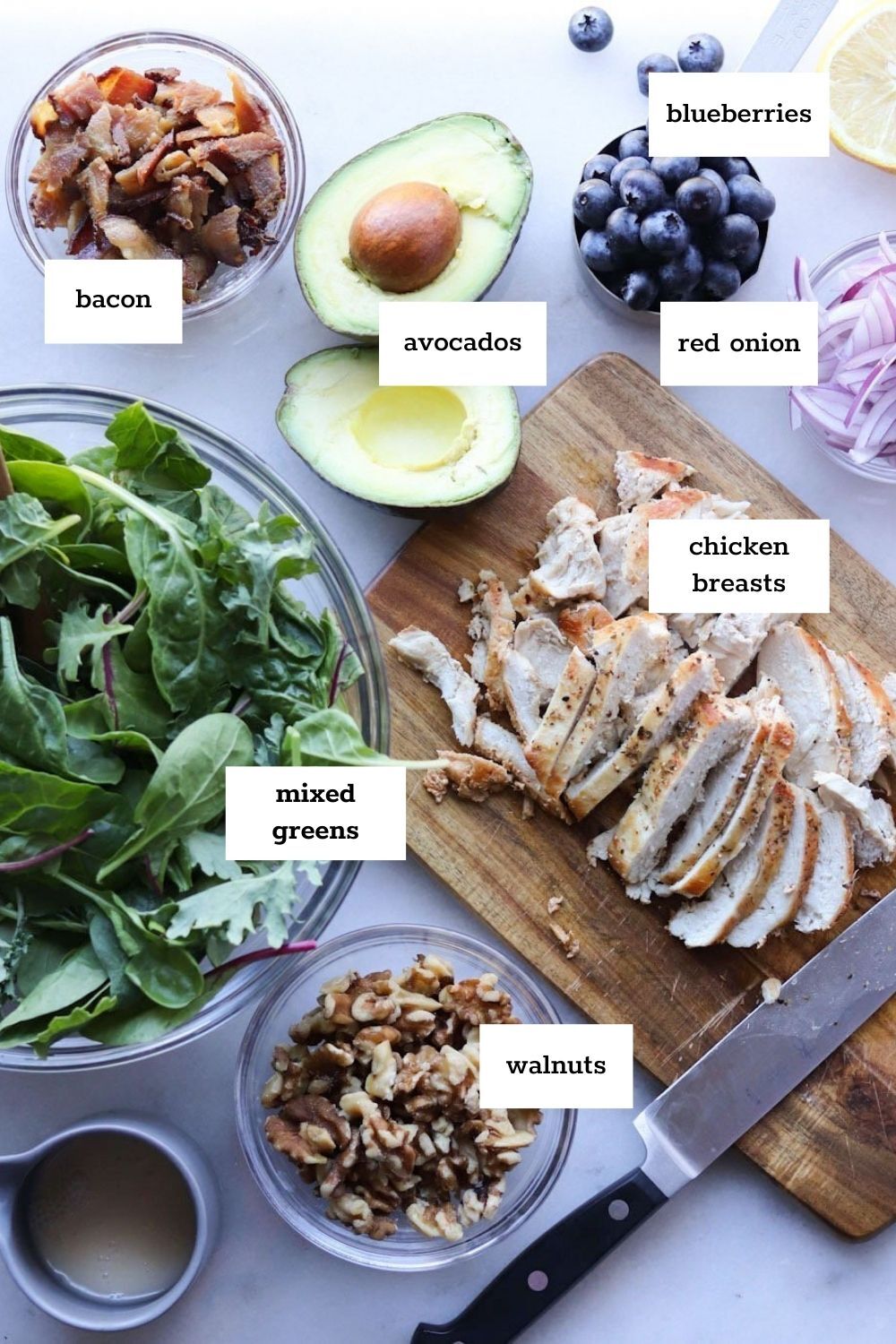 Sliced chicken breasts, avocados, red onion, blueberries, bacon, mixed greens, and walnuts are arranged on a table for a delicious high protein chicken salad meal prep.