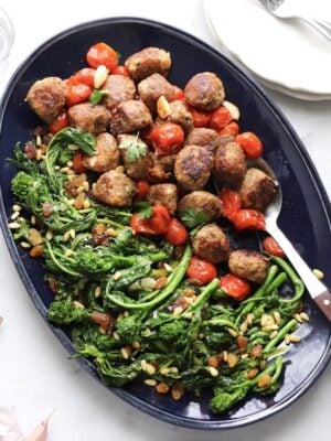 A long blue serving tray filled with meatballs, burst cherry tomatoes and broccoli rabe.