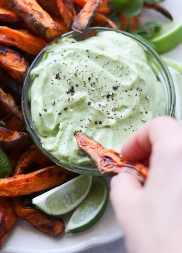A hand dips an Air Fryer Sweet Potato Fry into a bowl of creamy green dip, surrounded by more fries and lime wedges on a white plate.