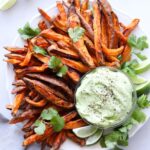 Cooked crispy sweet potato fries arranged on a white plate with a small glass dish of avocado aioli. The fries are sprinkled with black pepper and cilantro leaves and there are a few lime slices arranged on the plate.