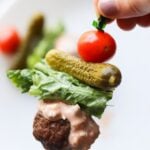 A hand holds a toothpick with a cherry tomato, mini pickle, lettuce, and mini burger bite.