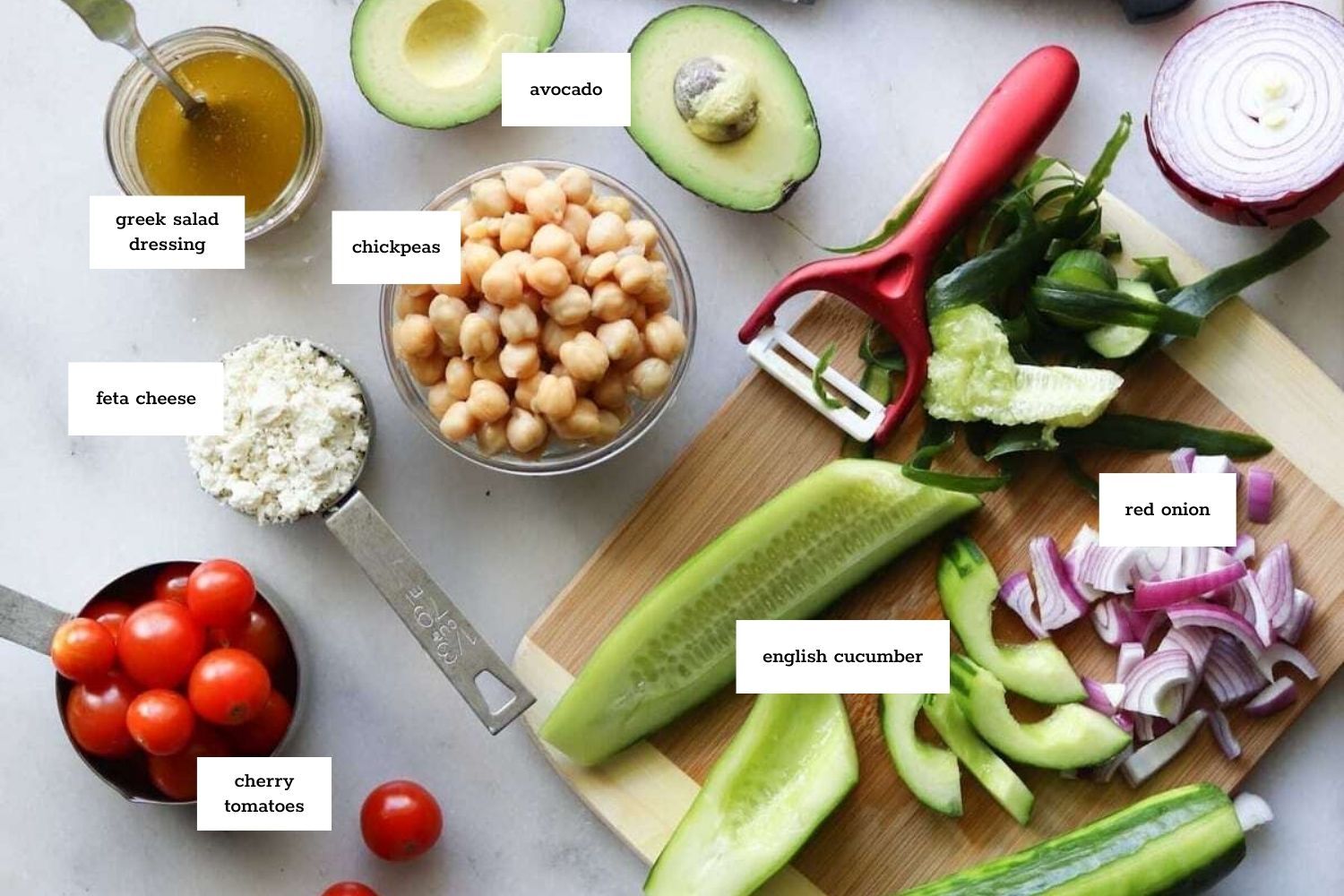 Top-down view of labeled ingredients for a chickpea avocado cucumber salad, including feta cheese, cherry tomatoes, green pepper, red onion, and Greek salad dressing.