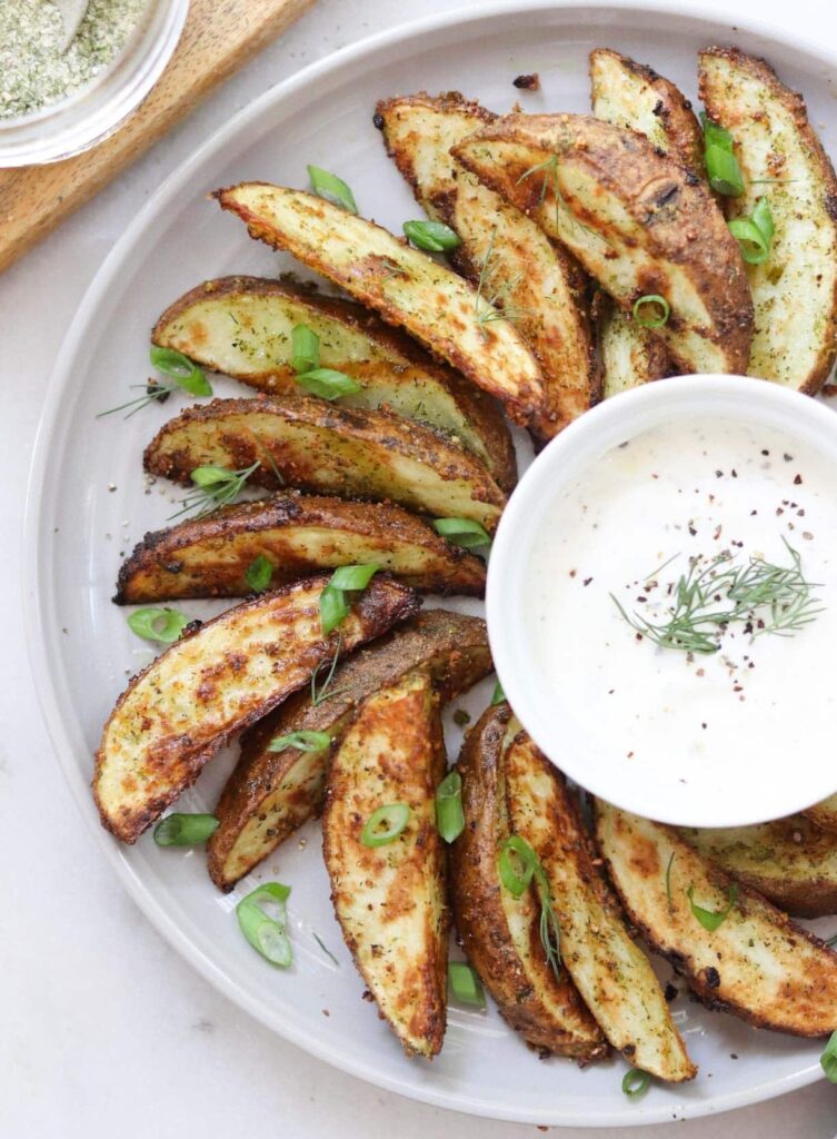 Crispy air fryer potato wedges arranged on a gray plate with a small dish of ranch, sprinkled with sliced green onions.