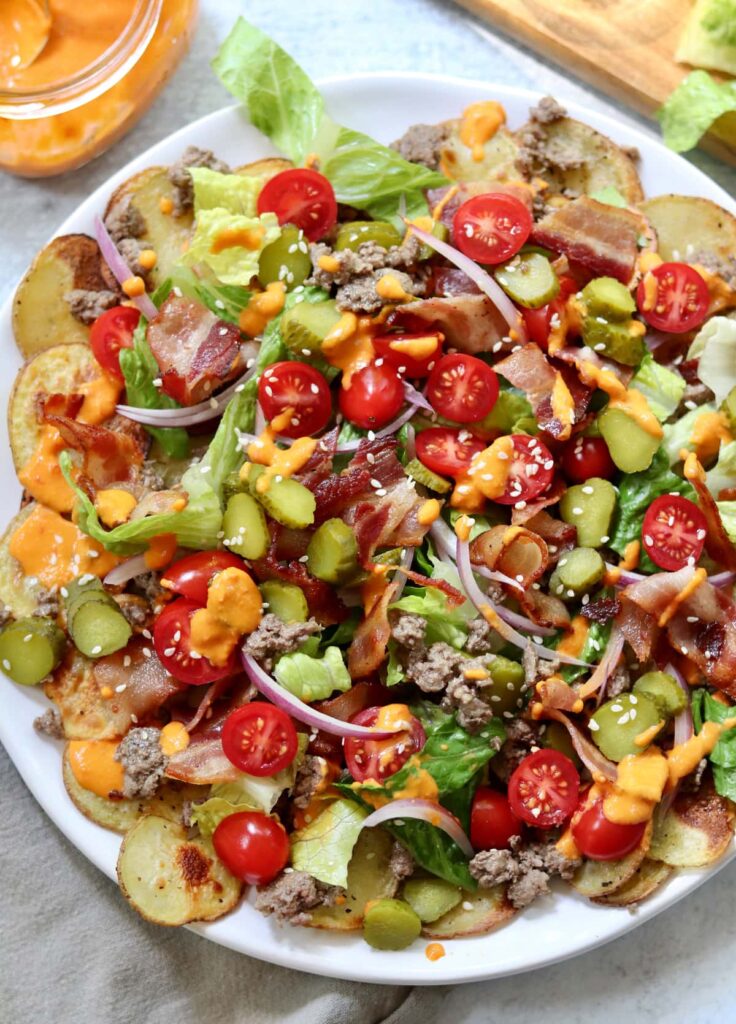 Large white plate filled with roasted potato rounds topped with ground beef, bacon, tomatoes, onion, and bacon and drizzled with red pepper "cheese" sauce.