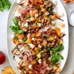 Top down of a large white platter on a white marble board filled with 5 grilled chicken breasts topped with diced apriums, mozzarella pears, sliced basil, and drizzled balsamic vinegar.