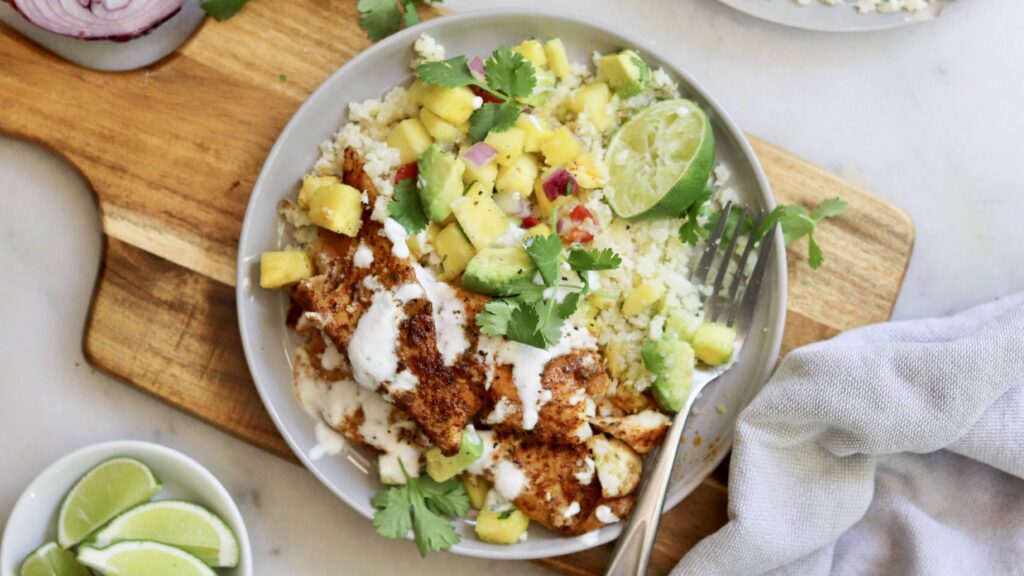 A plate of seasoned chicken with white sauce, rice, pineapple salsa, cilantro, and lime is served on a wooden board with extra lime wedges on the side—perfect alongside our signature blackened flounder for a fresh and vibrant meal.