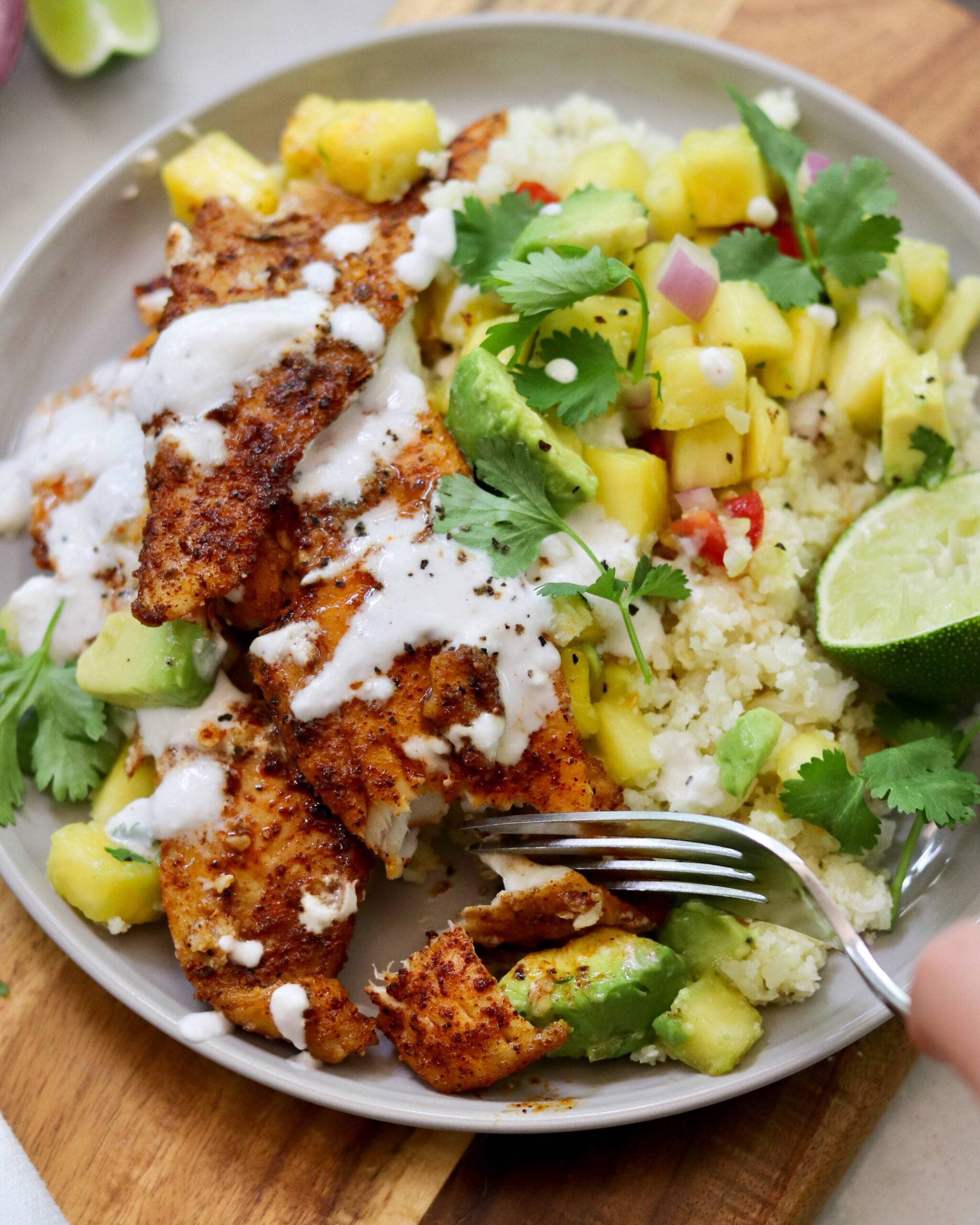 A plate with pan fried flounder, cauliflower rice, pineapple salsa, avocado slices, cilantro, and a drizzle of white sauce. A fork is held over the food.