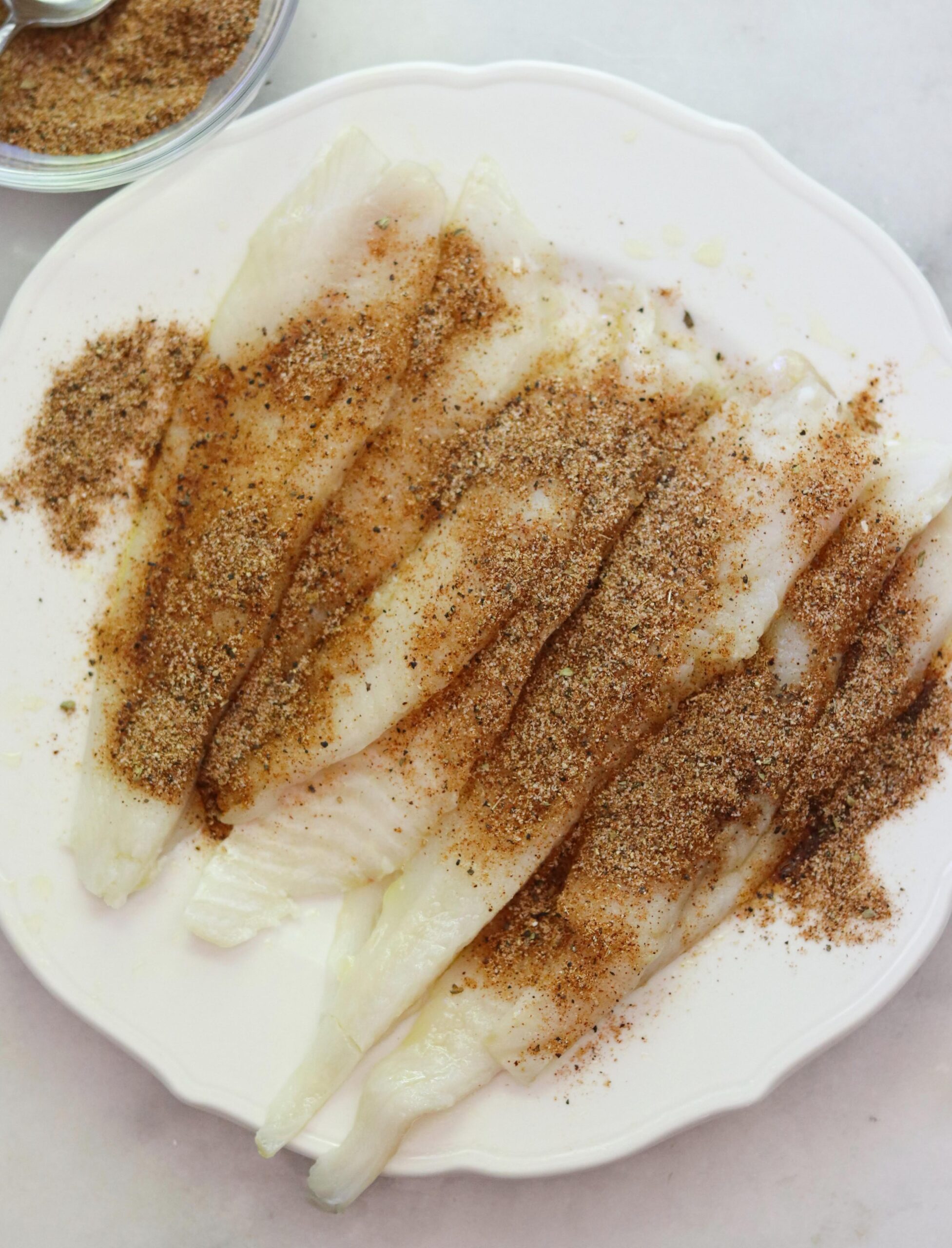 Raw flounder fillets arranged on a white plate, sprinkled generously with a brown spice mixture in preparation for blackened flounder. A small bowl with more spice mixture is visible in the corner.