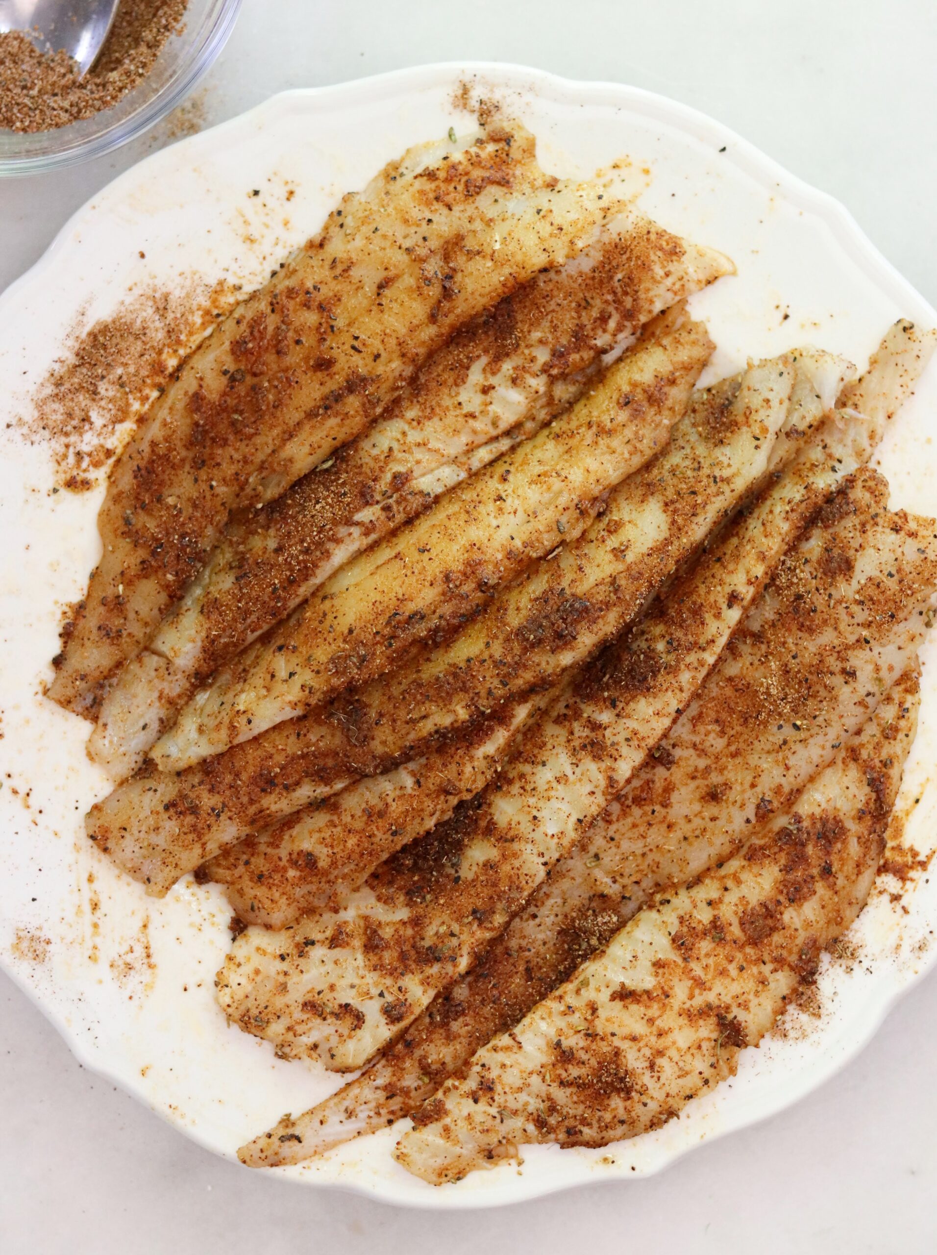 Raw fish fillets arranged on a white plate, coated with a reddish-brown spice blend reminiscent of classic blackened flounder, with extra seasoning visible in a small bowl nearby.