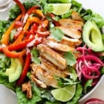 A large bowl filled with romaine lettuce, sliced chicken, pickled onions, cooked bell peppers, and garnished with slices of lime and cilantro.