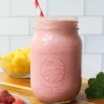 Side view close up of a mason jar filled with pink smoothie with a straw and two small dishes filled with strawberries and pineapples.