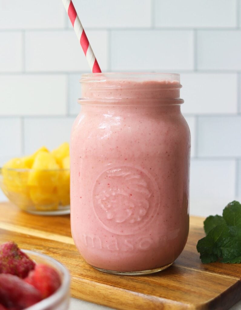 Side view close up of a mason jar filled with pink smoothie with a straw and two small dishes filled with strawberries and pineapples.