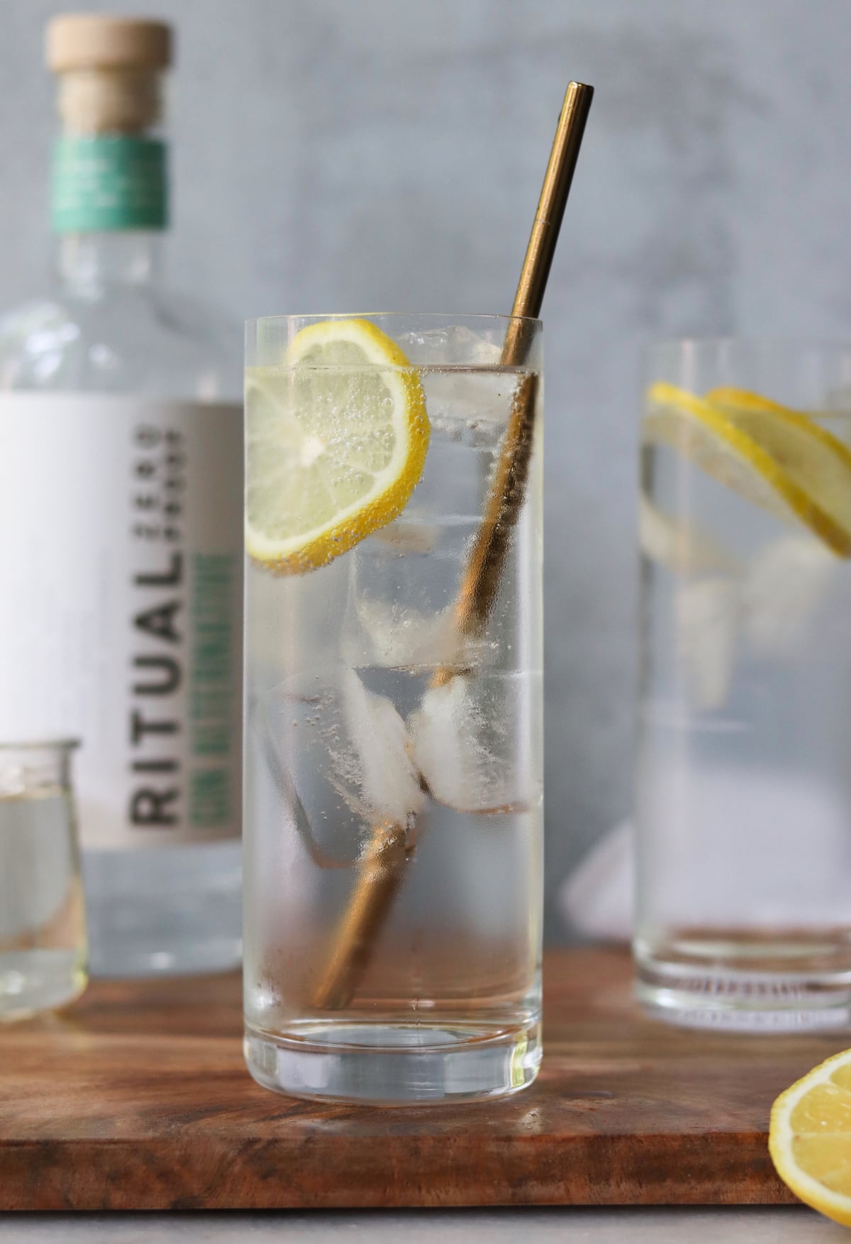 Side view close up of a tall, frosty glass filled with ice cubes and a clear drink. A lemon slice and gold straw are in the glass.