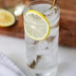 Close up of a tall glass filled with large ice cubes, a clear Tom Collins mocktail, garnished with a slice of lemon.