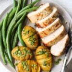 Close up top down of the plated pork loin, sliced, four mini hasselback potatoes topped with bright green gremolata, all beside steamed green beans.
