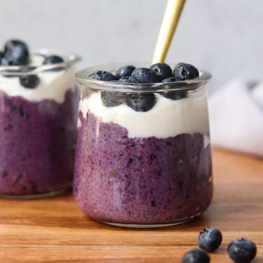 Two glass jars with blueberry chia pudding layered beneath yogurt and fresh blueberries. A golden spoon graces one jar, while a few scattered blueberries dot the wooden surface, completing this delightful scene.