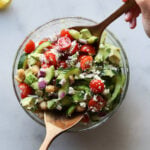 Salad tongs removing chickpea cucumber salad from a large glass salad bowl, viewed from overhead.