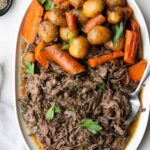 Top down of the shredded pot roast on a large white platter with lots of carrots, potatoes, and gravy.