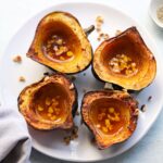 Top down of four roasted acorn squash halves on a white plate filled with melted butter and ghee and sprinkled with chopped hazelnuts.