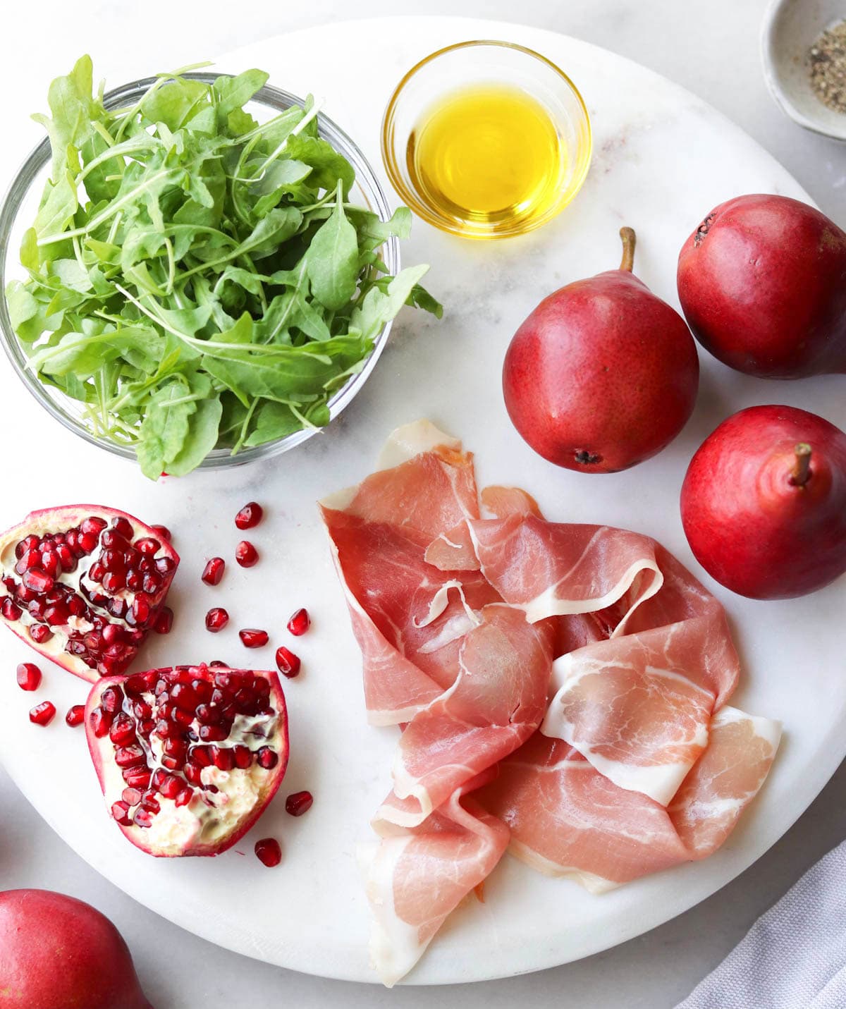 Recipe ingredients laid on a white marble board: arugula, oil, pomegranate, red pears, and prosciutto. 