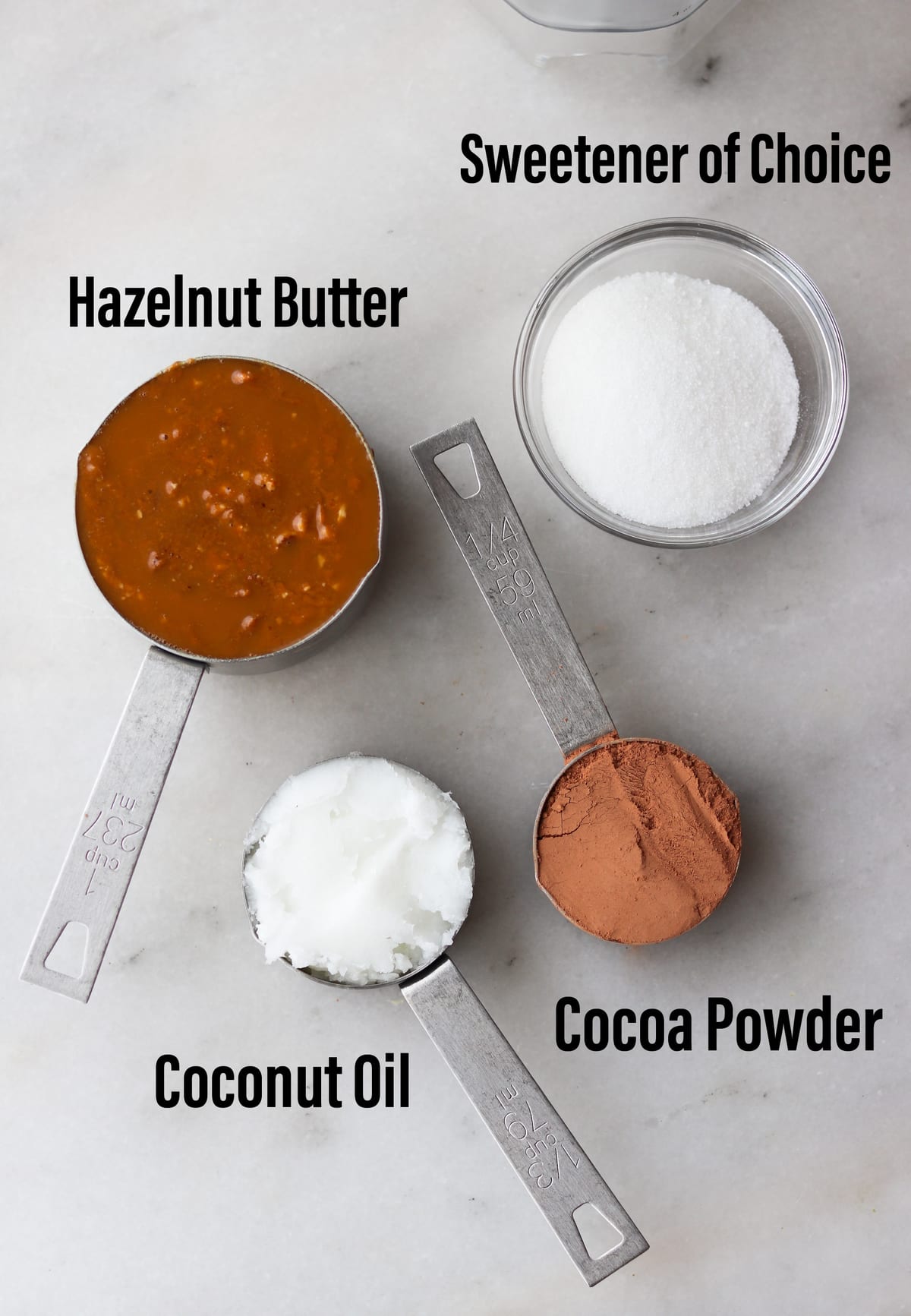 Top down of the recipe ingredients laid out on a marble board, labeled: hazelnut butter, sweetener, coconut oil, and cocoa powder.
