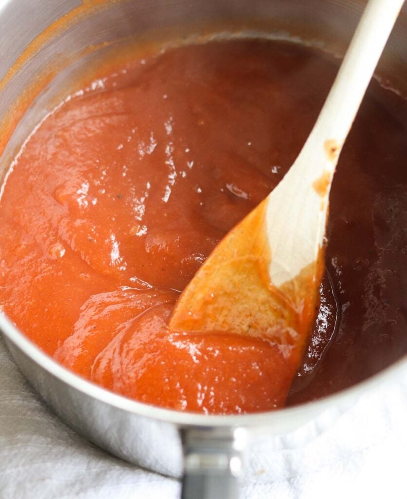 A wooden spoon stirs a thick, reddish-orange sugar free barbecue sauce in a stainless steel saucepan.