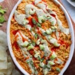 Top down of the cooked buffalo chicken dip in an oval dish, topped with hot sauce, ranch, diced celery, and green onions.