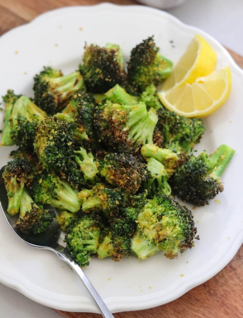 Overhead view of air fried broccoli florets on a white plate with a serving spoon and a lemon wedge.