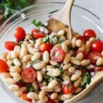Cannellini bean salad in a glass bowl with wooden serving spoons.