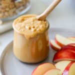 Side view of peanut butter in a small glass jar beside slices of apples.