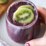 Top down of a hand holding a large glass filled with dark purple kiwi spinach and blueberry smoothie, topped with two slices of kiwi.