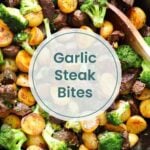 A skillet filled with cooked steak bites, halved baby potatoes, and broccoli florets. A wooden spoon rests in the pan. Text overlay reads "Garlic Steak Bites." Perfect for keto lovers, just like strawberry cheesecake fat bombs.