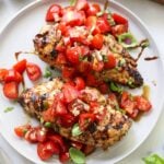 Two plated grilled chicken breasts topped with bright red tomato bruschetta, basil, and vinegar.