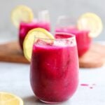 Side view of three glasses filled with bright pink mango dragonfruit lemonade, garnished with slices of lemon.